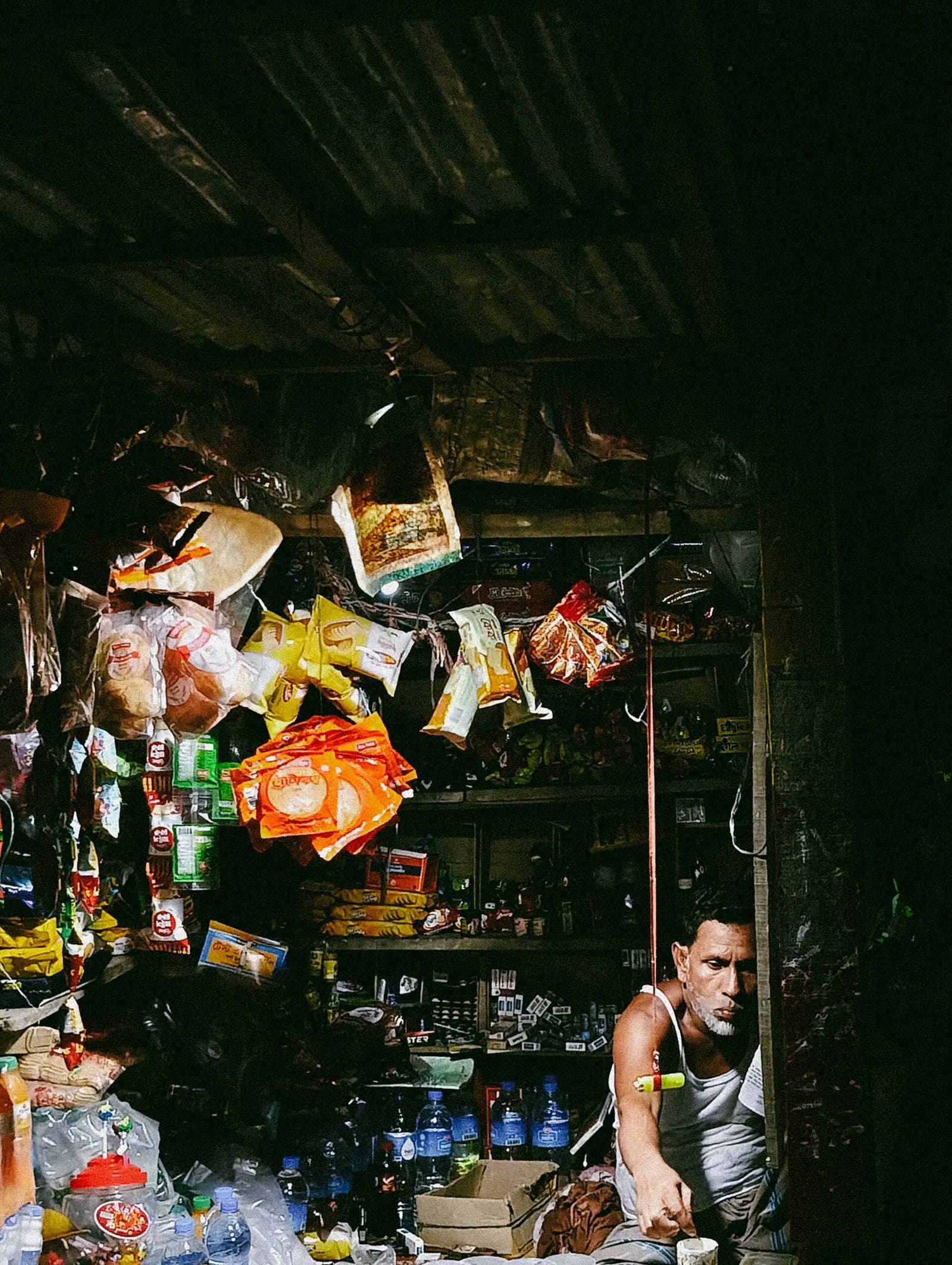 A shopkeeper sitting in his small shop in Bangladesh, surrounded by hanging packets of snacks and shelves of goods.
