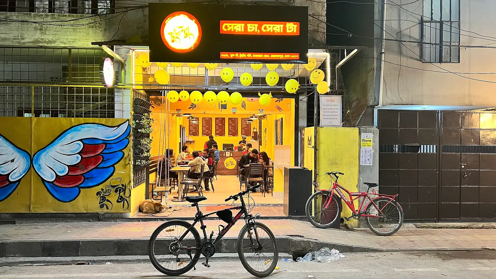 A brightly lit tea shop on a Dhaka street at night, customers sitting at tables inside.