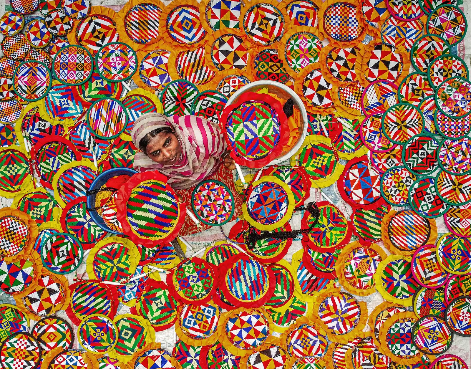 Overhead view of a woman surrounded by dozens of brightly patterned handmade fans she has stitched.