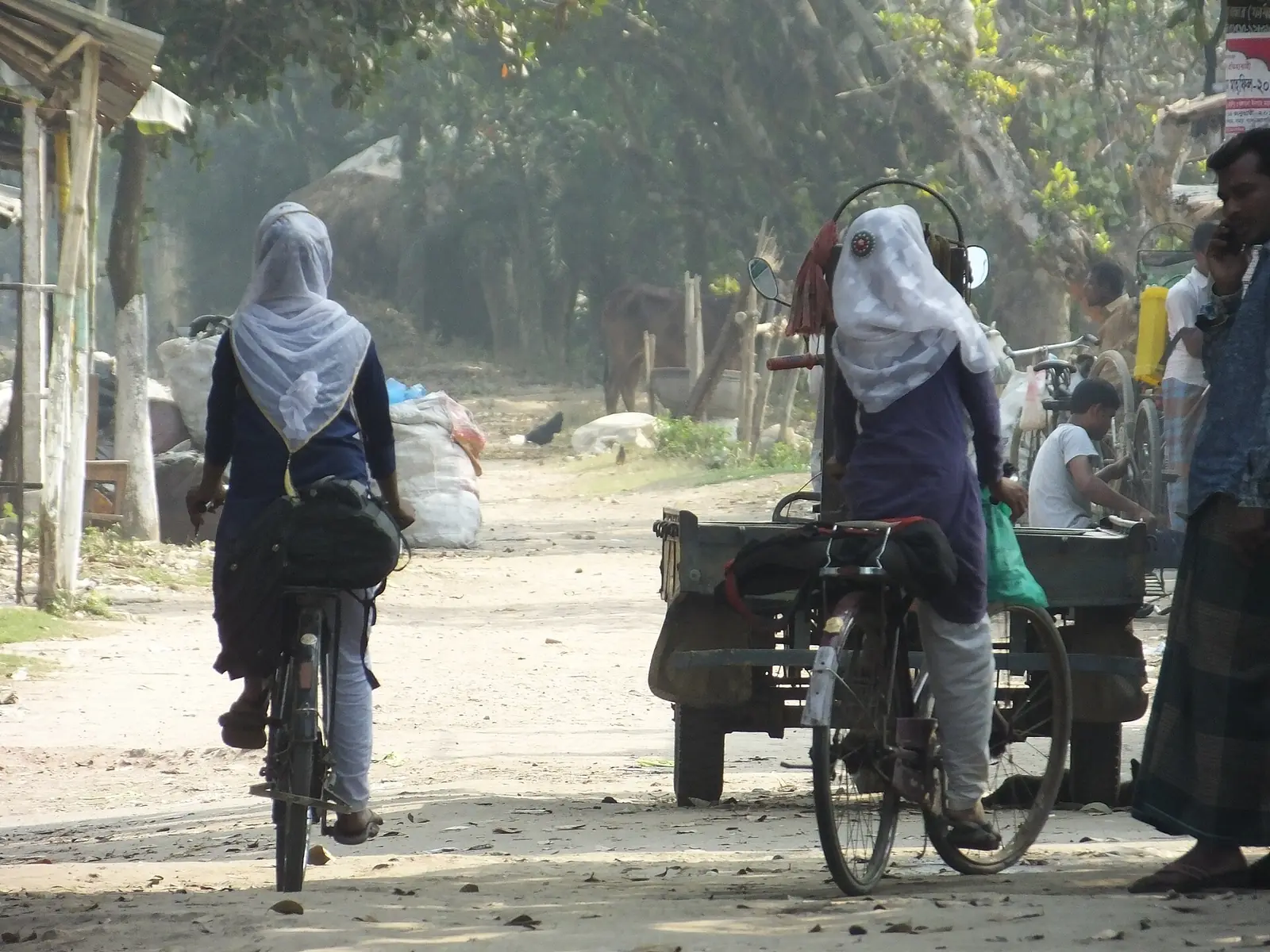 Two schoolgirls in uniform cycling along a dirt village road in Bangladesh on their way to school.
