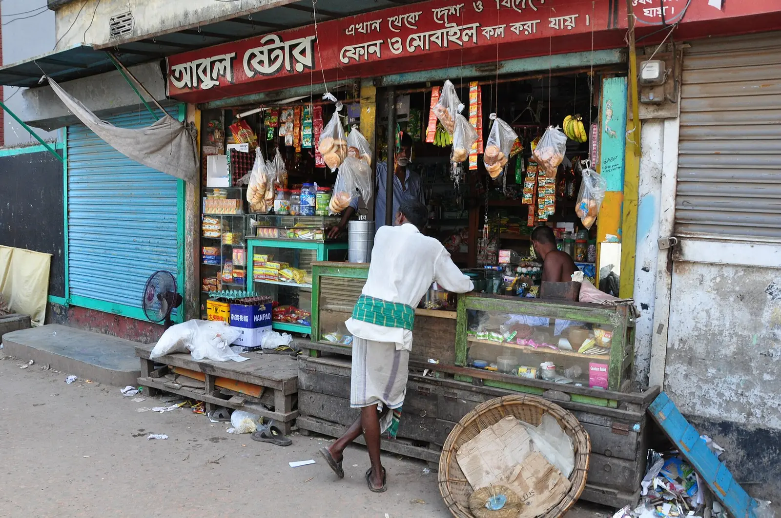 A neighbourhood grocery shop in Keraniganj, Bangladesh, its Bengali signboard offering phone and mobile calls, with a customer leaning on the counter.